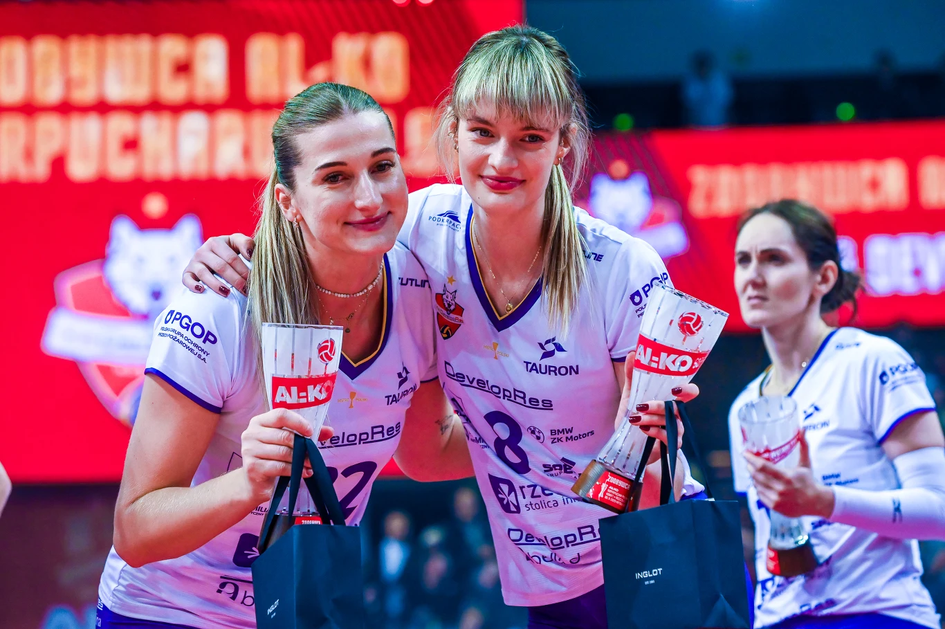 Dwie zawodniczki siatkarskie w białych koszulkach drużynowych stoją obok siebie z uśmiechami, trzymając puchary i torby z nagrodami, w tle widoczna jest trzecia siatkarka oraz jasne światła areny sportowej.