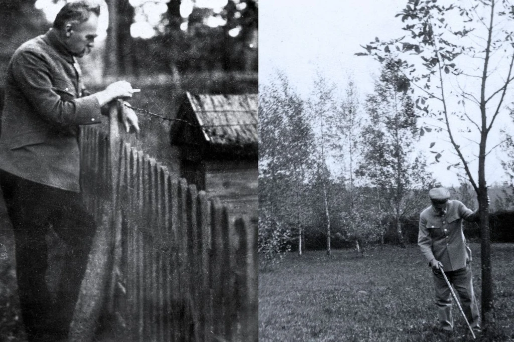 Mężczyzna w średnim wieku w marynarce spędza czas na podwórzu, z lewej strony oparty o drewniany płot i obserwuje ogród, z prawej wykonuje pracę ogrodową przy młodym drzewie; w tle niskie zabudowania i rząd drzew.