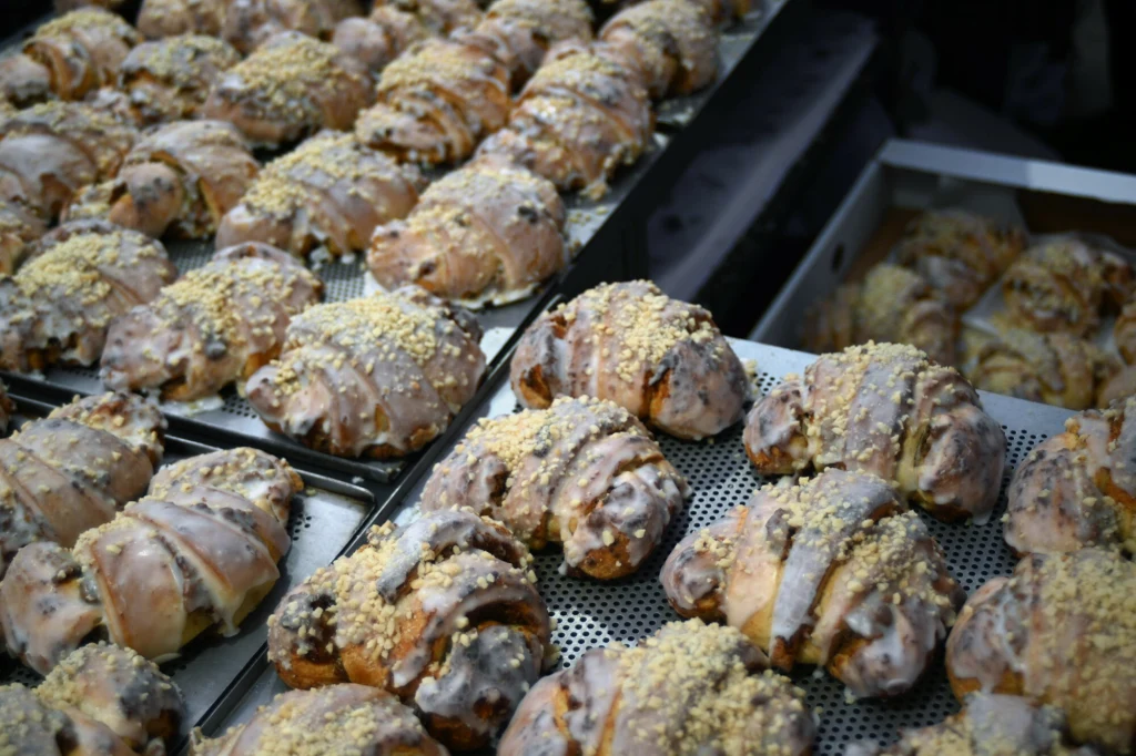 Rzędy polukrowanych i obsypanych kruszonką półksiężyców z ciasta drożdżowego, ułożonych na blachach, sugerujących przygotowanie do sprzedaży lub podania.