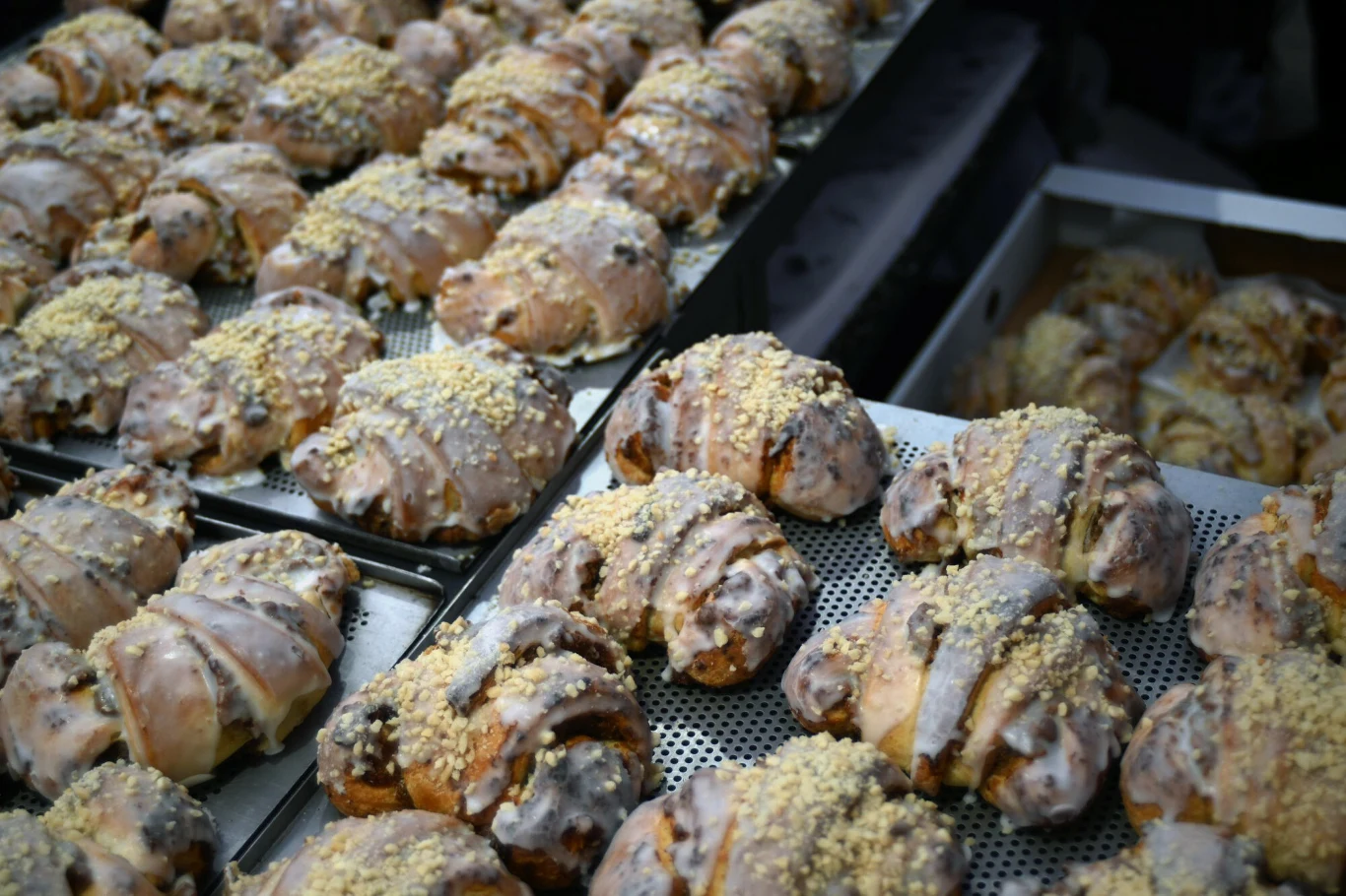 Rzędy polukrowanych i obsypanych kruszonką półksiężyców z ciasta drożdżowego, ułożonych na blachach, sugerujących przygotowanie do sprzedaży lub podania.