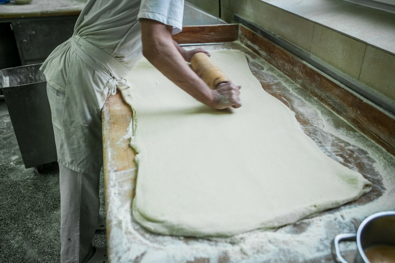 Osoba wałkuje duży kawałek ciasta na obsypanym mąką stole w profesjonalnej kuchni lub piekarni, widoczne ręce uformowane nad wałkiem oraz otoczenie z blatem do wypieku.