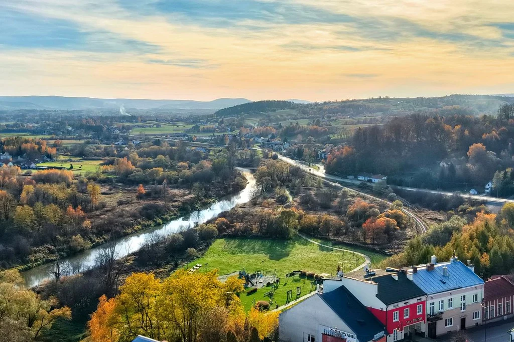 Malownicza dolina z wijącą się rzeką, otoczona polami, lasami i wzgórzami, na pierwszym planie zabudowania z kolorowymi dachami, jasne niebo pokryte delikatnymi chmurami oraz jesienna roślinność tworząca spokojny, sielski nastrój.