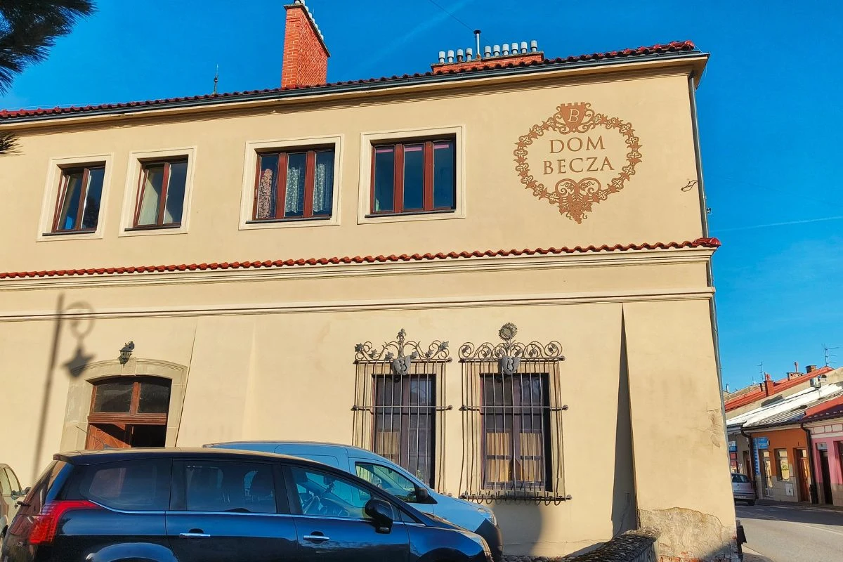 Dwupiętrowy, kremowy budynek z czerwonym dachem, ozdobnym napisem 'Dom Becz'a i dekoracją na fasadzie. Przy budynku zaparkowany czarny samochód. Po prawej stronie widać fragment ulicy i sąsiednich domów.