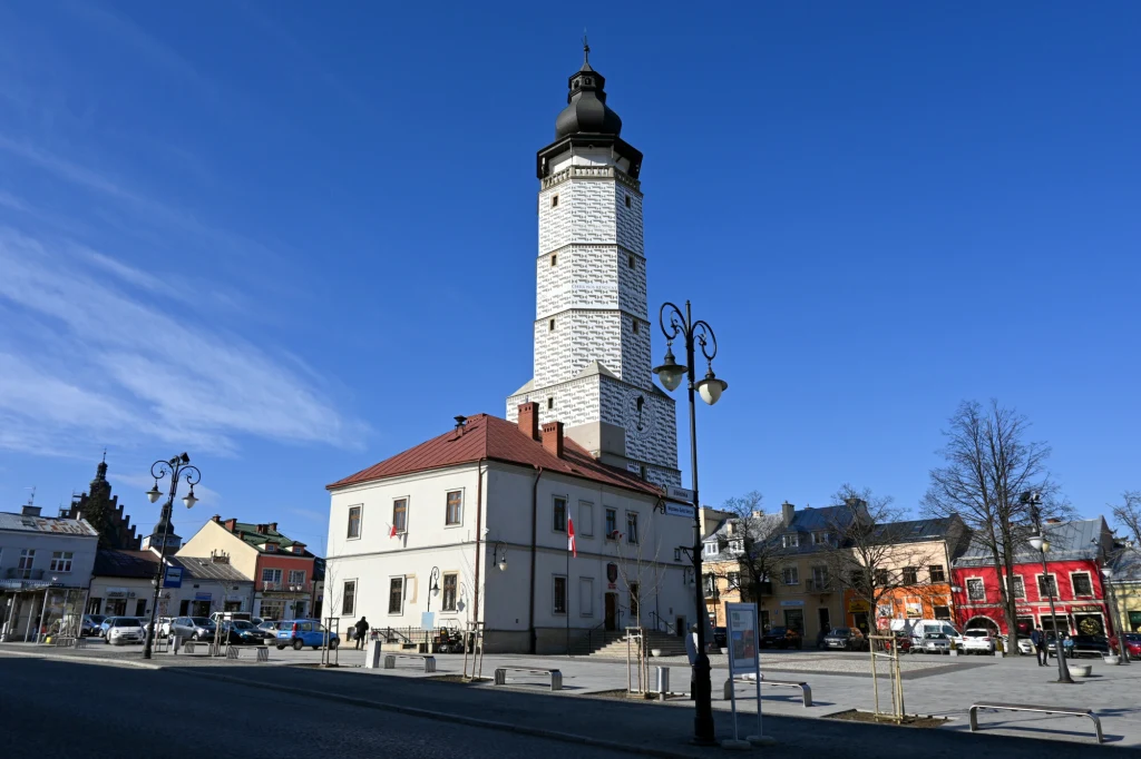 Historyczny ratusz z wysoką wieżą dominuje nad pustym rynkiem otoczonym przez kolorowe kamienice. Przed budynkiem znajdują się latarnie i zaparkowane samochody, a po prawej stronie widoczne są drzewa bez liści.