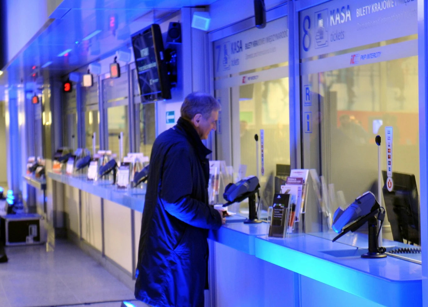 Za 18-minutową podróż pociągiem z centrum Krakowa na lotnisko w Balicach pasażerowie płacą 20 złotych Mężczyzna w średnim wieku w czarnym płaszczu stoi przy okienku kasowym w nowoczesnym, dobrze oświetlonym holu, otoczony szeregiem podobnych stanowisk kasowych z monitorami i informacjami o biletach.