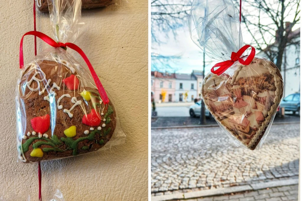 Pierniki w kształcie serca ozdobione kolorowym lukrem, jeden z napisem oraz motywami kwiatów, oba zapakowane w przezroczystą folię i przewiązane czerwoną wstążką, tło stanowi fragment ulicy z brukowaną nawierzchnią i zabudowaniami.