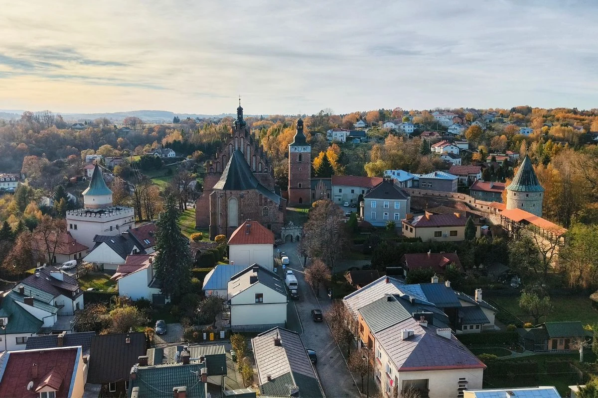 Panorama miasta z widoczną zabytkową architekturą, w tym dużym gotyckim kościołem i wieżą, otoczona domami jednorodzinnymi oraz bujną roślinnością. W tle wzgórza i jesienne drzewa tworzą malowniczy krajobraz.