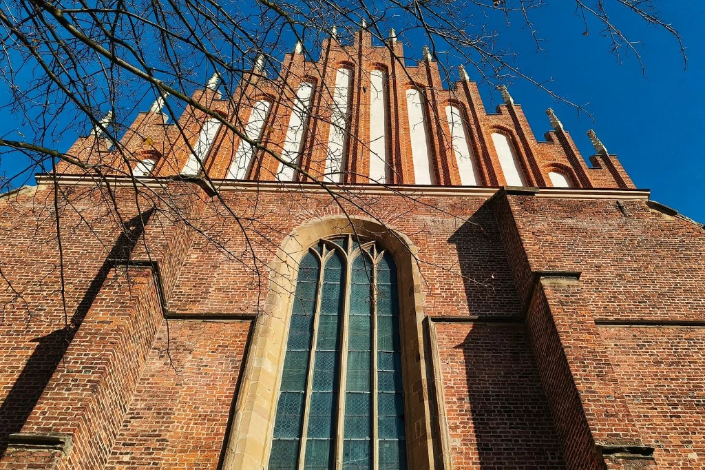 Fasada gotyckiego kościoła z czerwonej cegły z wysokimi ostrołukowymi oknami, skierowana w górę pod błękitnym niebem, ozdobiona pionowymi przyporami i strzelistymi zdobieniami.