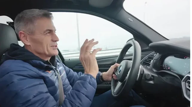 Mężczyzna w średnim wieku ubrany w niebieską kurtkę siedzi za kierownicą samochodu, trzymając ręce na kierownicy i gestykulując, w tle widoczne rozmyte okno i zarys jezdni.