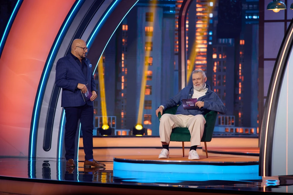 Dwóch mężczyzn w studiu telewizyjnym, jeden siedzi wygodnie na zielonym fotelu trzymając kartkę, drugi stoi obok w eleganckim garniturze, w tle kolorowe światła i nowoczesna scenografia.