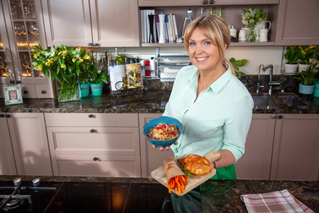 Uśmiechnięta kobieta stojąca w nowoczesnej kuchni, trzymająca w rękach miseczkę sałatki oraz deskę z pieczywem i świeżymi warzywami. W tle eleganckie szafki kuchenne, kwiaty w wazonach i zioła doniczkowe.