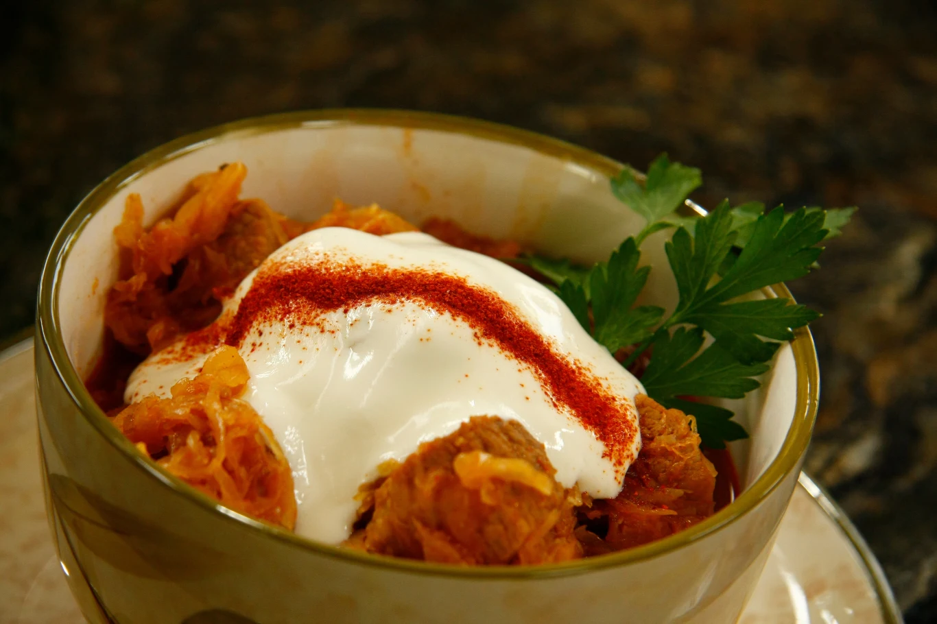 Gulasz segedyński Porcja bigosu z mięsem, udekorowana śmietaną i czerwoną papryką, obok zielona natka pietruszki.