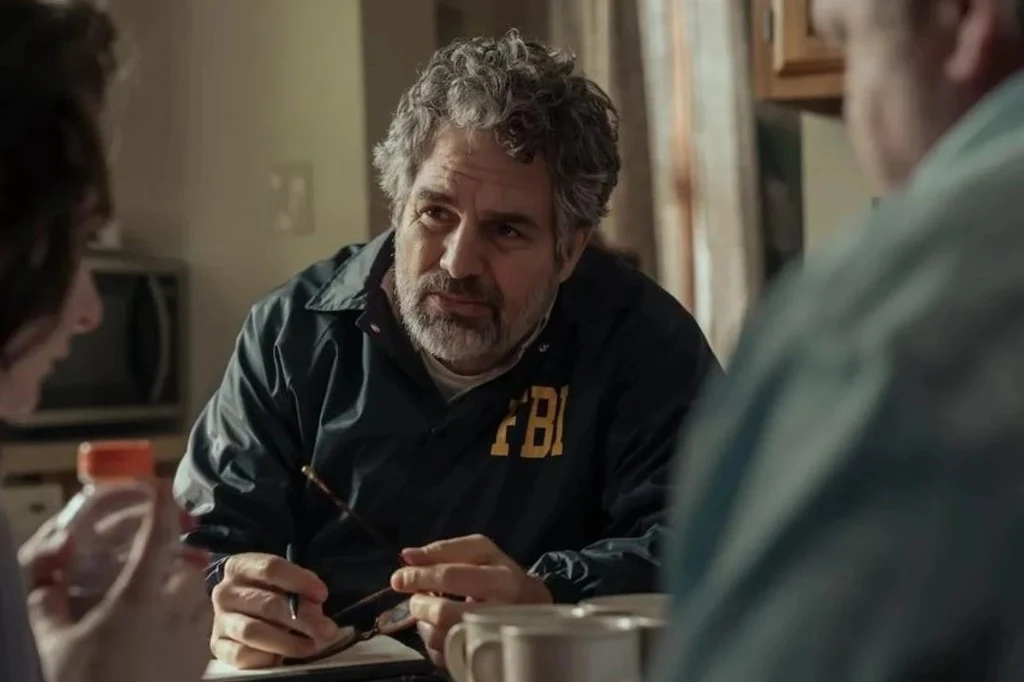 Mężczyzna z siwiejącymi włosami w kurtce z napisem FBI prowadzi rozmowę przy stole z dwiema innymi osobami, w tle kuchnia oraz kubki na stole.