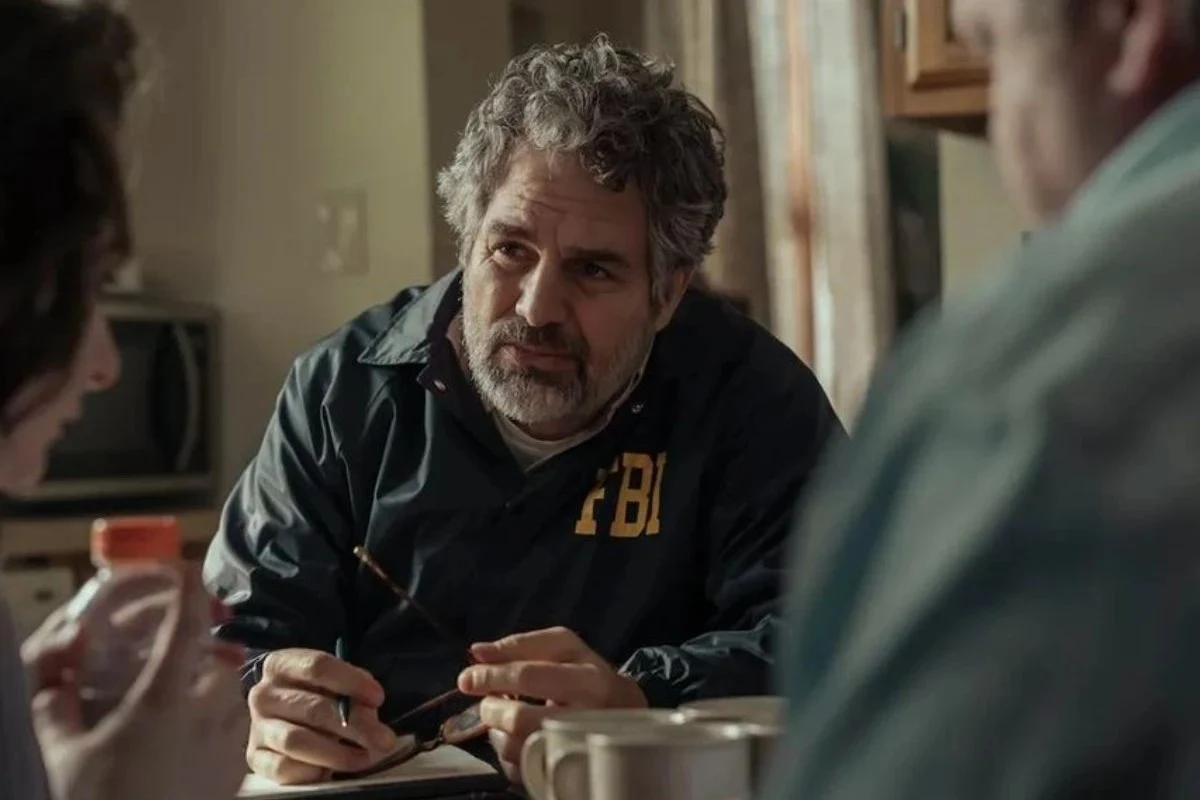Mężczyzna z siwiejącymi włosami w kurtce z napisem FBI prowadzi rozmowę przy stole z dwiema innymi osobami, w tle kuchnia oraz kubki na stole.