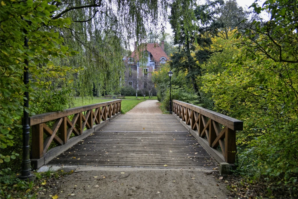 Ogromny park i aboretum zachęcają do spacerów i podziwiania zamku z różnych perspektyw Drewniany mostek prowadzący przez park otoczony bujną zielenią, w tle widoczny jest elegancki budynek z czerwonym dachem, częściowo przesłonięty przez drzewa.