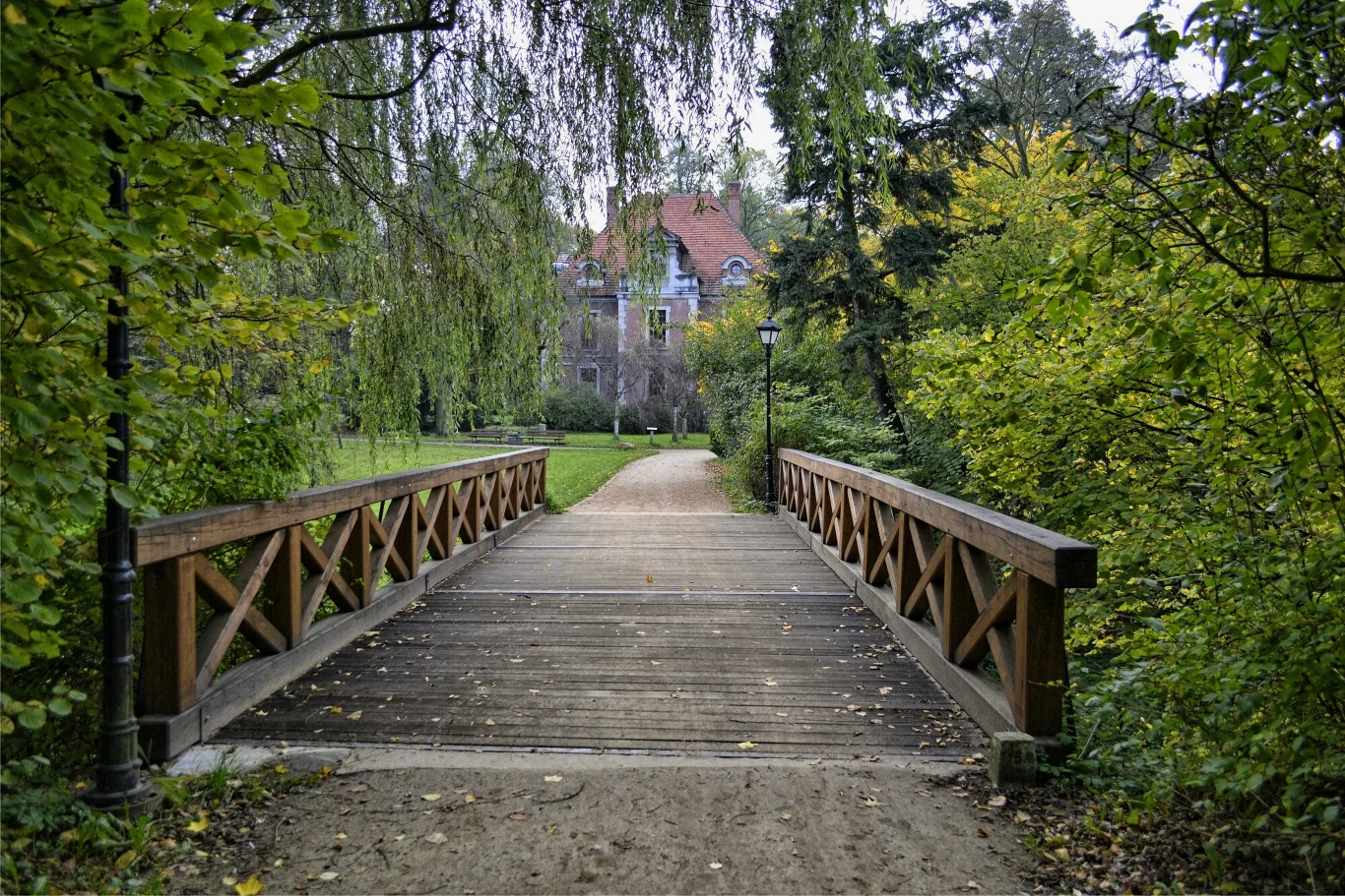 Ogromny park i aboretum zachęcają do spacerów i podziwiania zamku z różnych perspektyw Drewniany mostek prowadzący przez park otoczony bujną zielenią, w tle widoczny jest elegancki budynek z czerwonym dachem, częściowo przesłonięty przez drzewa.