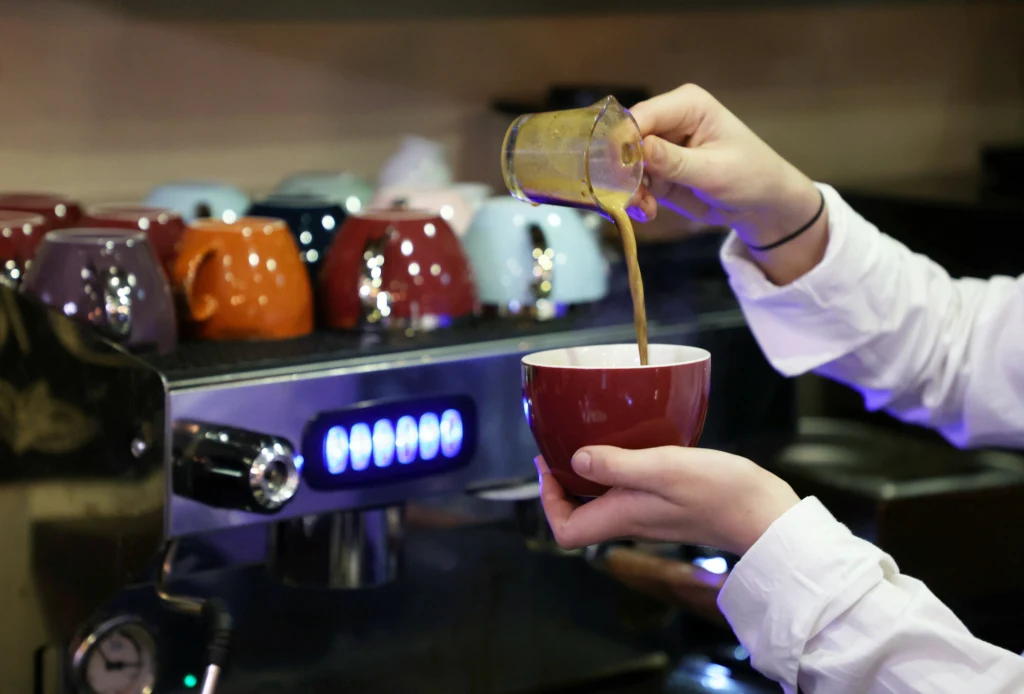 Mimo wysokich cen Polacy nie rezygnują z kawy na mieście Barista wlewa świeżo zaparzoną kawę z dzbanuszka do czerwonej filiżanki stojącej w dłoni, w tle widoczne są kolorowe filiżanki ułożone na ekspresie do kawy.