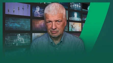 Doświadczony mężczyzna o siwych włosach na tle multimedialnej ściany z ekranami przedstawiającymi cyfrowe grafiki i postacie techniczne.