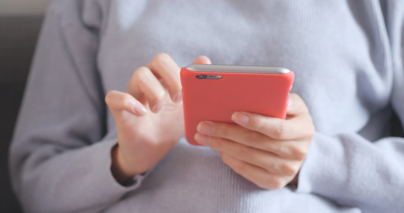 Osoba trzymająca smartfon w różowym etui, ubrana w jasną bluzę, wykonuje gest palcem na ekranie telefonu, siedząc w wygodnej pozycji.