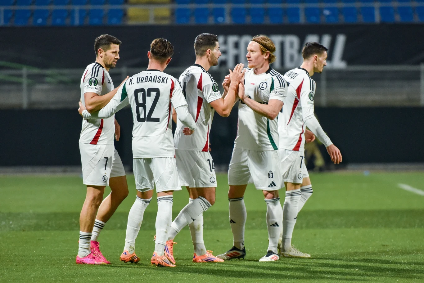 Piłkarze Legii Warszawa w sezonie 2025/2026 Grupa piłkarzy w białych strojach świętuje zdobycie gola na boisku, widoczne emocje i gesty zadowolenia, stadion w tle, jeden zawodnik z numerem 82, otoczenie sportowe.