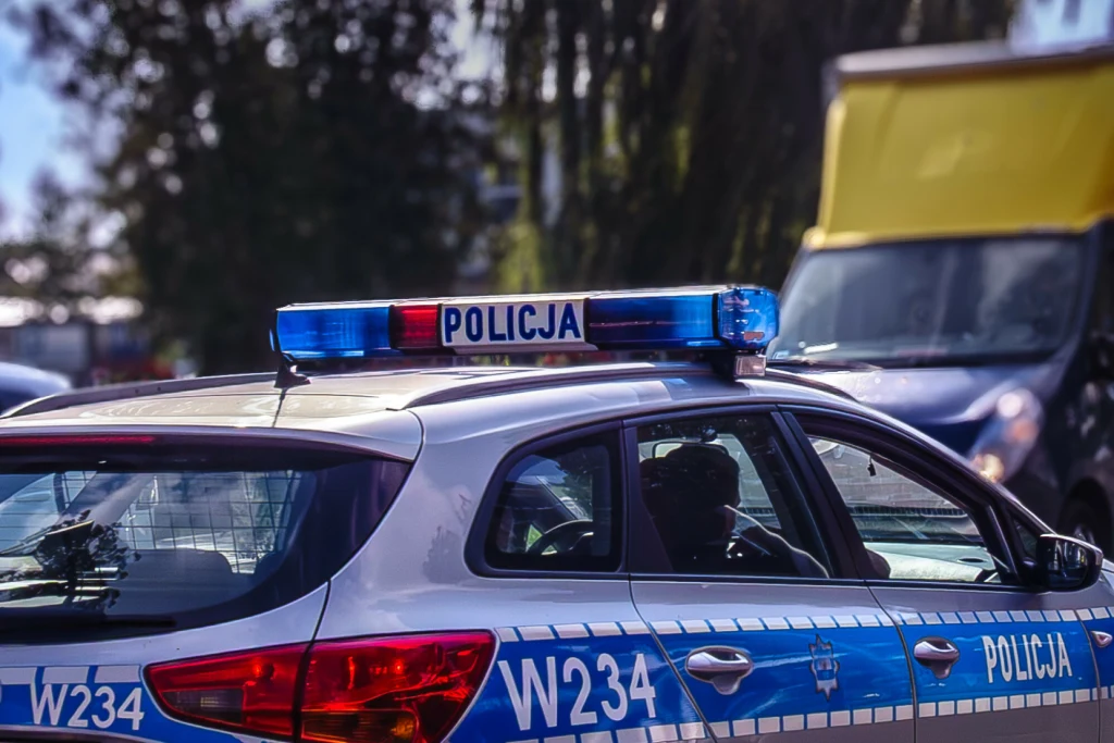 Sędzia zatrzymana w stanie nietrzeźwości. Prokuratura postawiła zarzut Radiowóz policyjny z widocznym oznaczeniem i sygnalizacją świetlną, stojący na ulicy obok innego samochodu. W tle drzewa i fragment żółtego auta dostawczego.