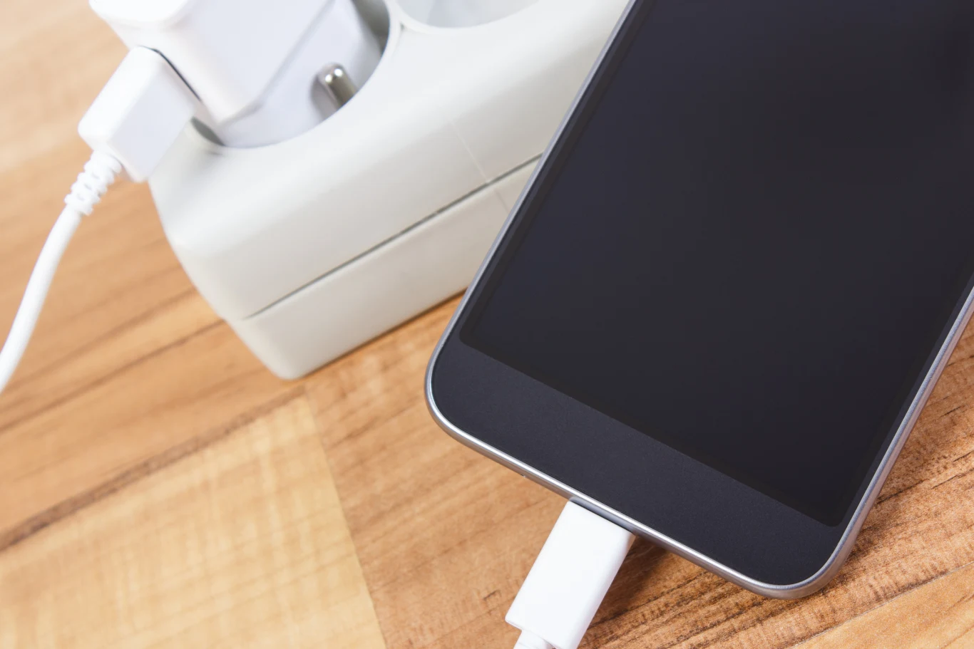 Smartfon ładowany za pomocą kabla USB podłączonego do zasilacza sieciowego, całość umieszczona na drewnianym blacie.