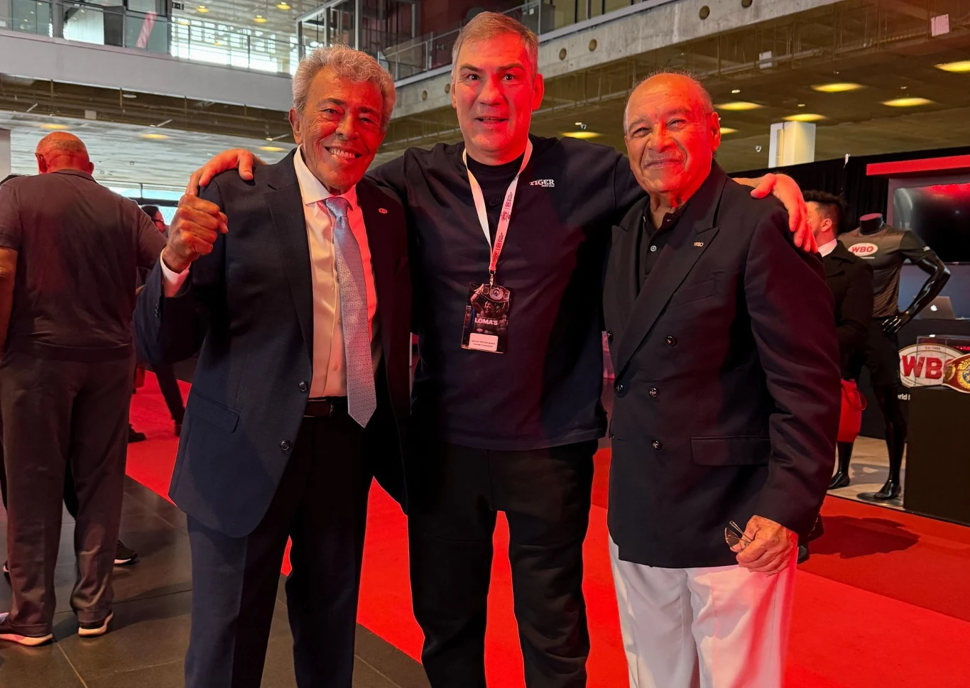 Trzech mężczyzn pozuje razem podczas wydarzenia w przestronnym, nowoczesnym wnętrzu z czerwonym dywanem, na jednym z nich zawieszony jest identyfikator, pozostali dwaj są ubrani elegancko i uśmiechają się.