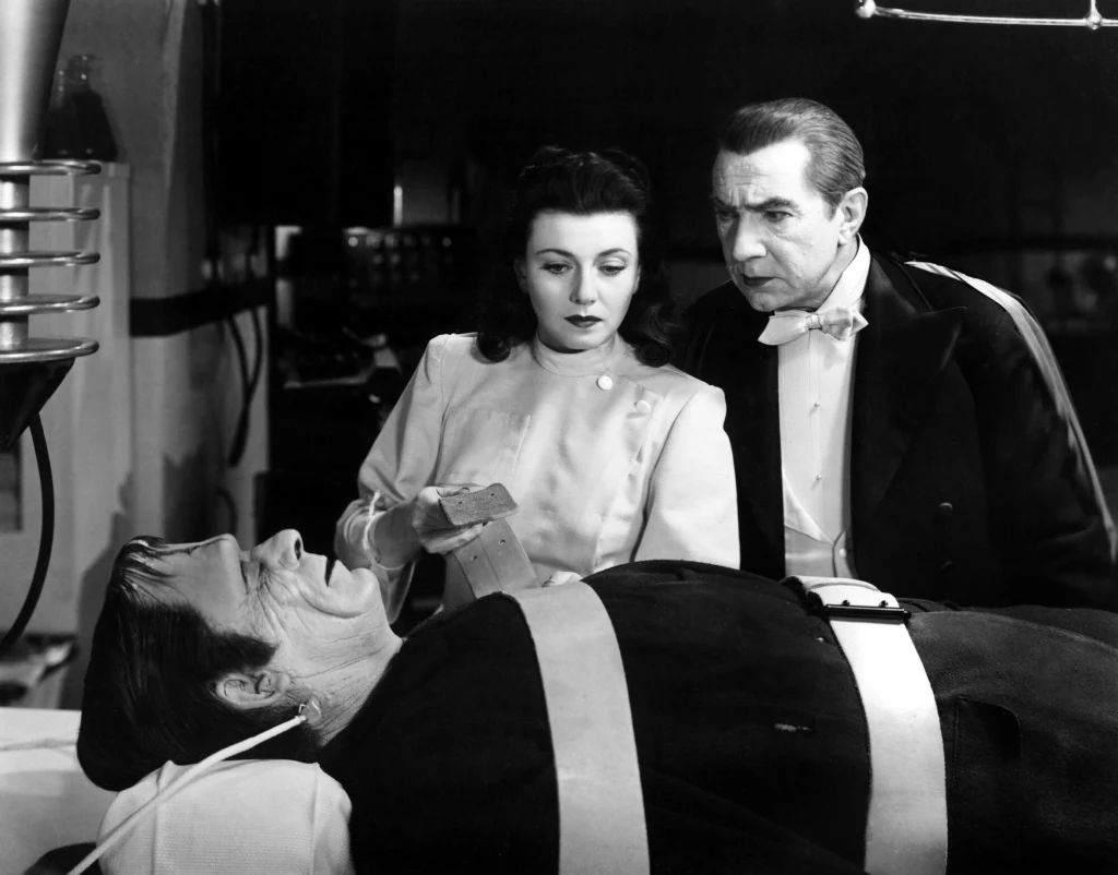 Dwóch ludzi stoi obok leżącego na stole mężczyzny, który jest unieruchomiony pasami, całość odbywa się w laboratorium w stylu retro, obecna jest także aparatura laboratoryjna i kartka trzymana przez kobietę.