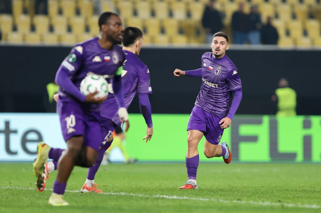 Afimico Pululu, Bernardo Vital i Sergio Lozano w barwach Jagiellonii Białystok Piłkarze w fioletowych strojach podczas rozgrywki na stadionie, jeden z nich trzyma piłkę, a pozostali są w ruchu, na trybunach widzowie, w tle rozmazane żółte krzesełka.