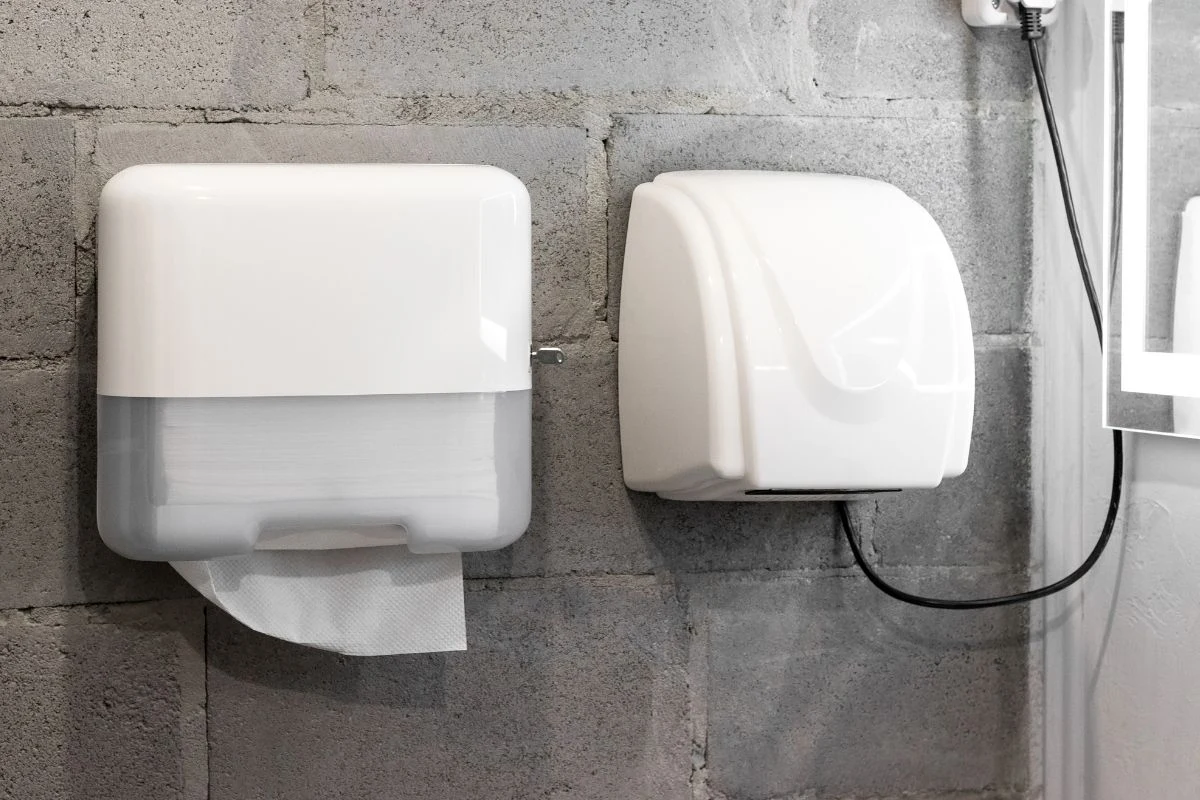 Dozownik ręczników papierowych oraz elektryczna suszarka do rąk zamontowane na szarej, surowej ścianie z betonu, obok siebie w łazience.