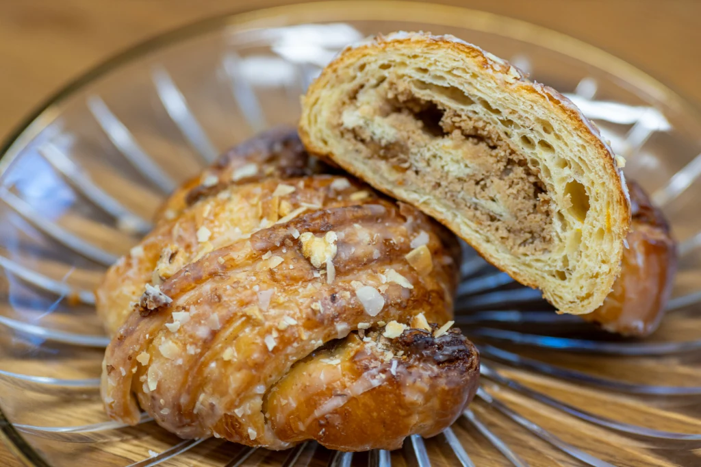 Rogal z ciasta drożdżowego pokryty lukrem i posypany orzechami, na talerzyku, z widocznym przekrojem ukazującym nadzienie orzechowe.