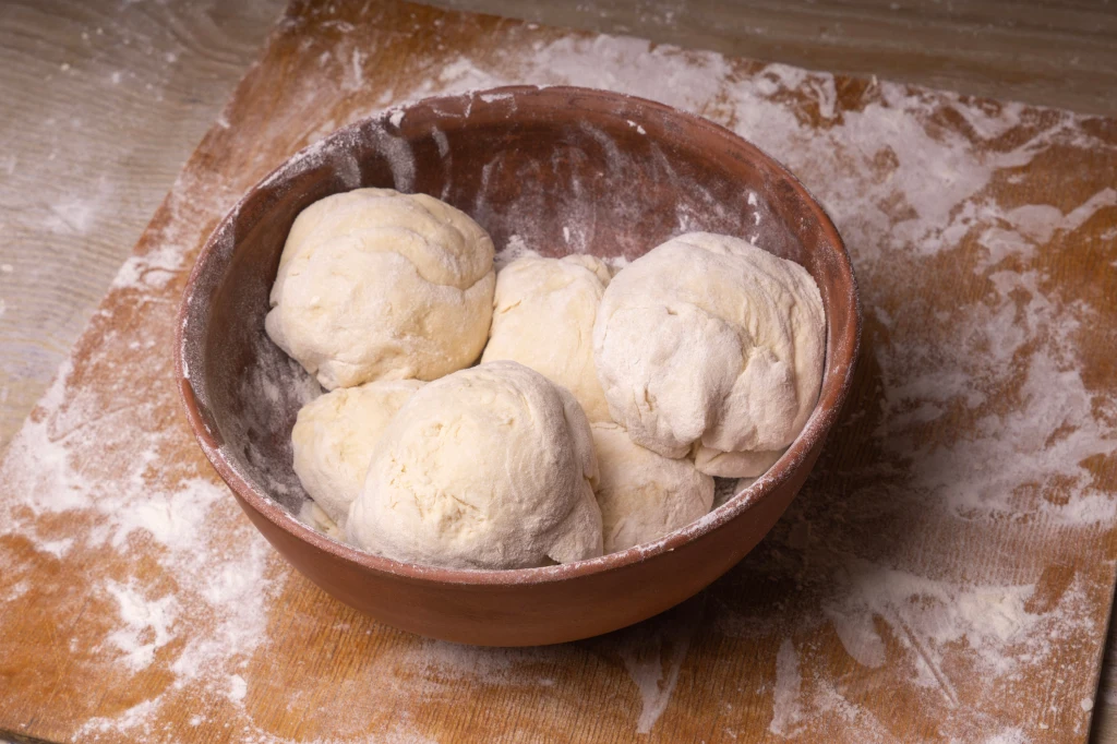 Mąka i co jeszcze? Oto pomysły na dodatki do ciasta pierogowego Kilka porcji ciasta uformowanych w kule leży w glinianej misce pokrytej mąką. Miska ustawiona jest na oprószonej mąką stolnicy.