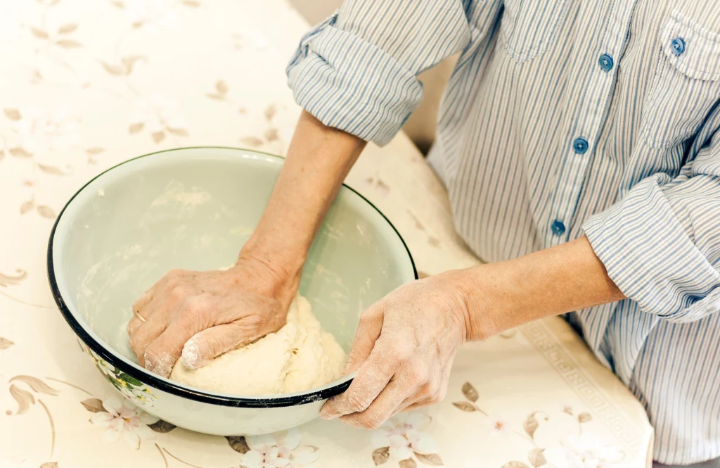 Ciasto na pierogi trzeba długo wyrabiać Ręce osoby wyrabiające ciasto w dużej, emaliowanej misce ustawionej na stole przykrytym jasnym obrusem w kwiaty.