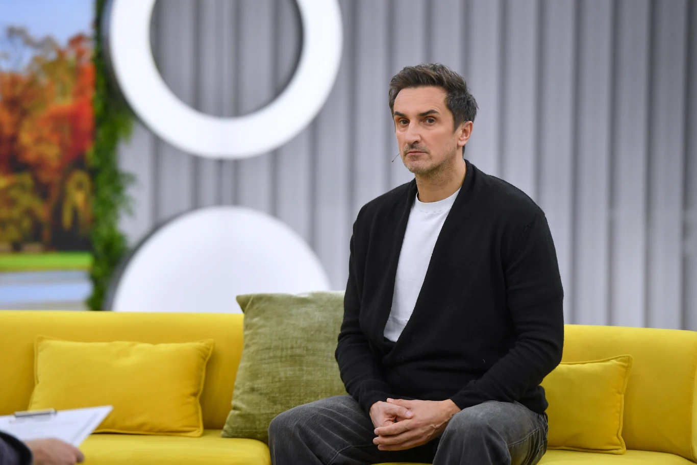 Sebastian Karpiel-Bułecka stoi na czele grupy Zakopower Mężczyzna o ciemnych włosach w czarnym swetrze i białej koszulce siedzi na żółtej sofie z zieloną poduszką na tle jasnych geometrycznych kształtów i szarej ściany.