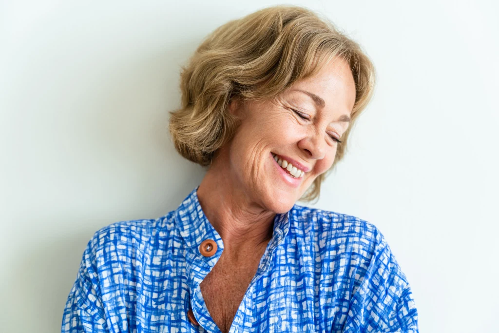 Starsza kobieta z jasnobrązowymi włosami uśmiecha się i odwraca głowę w bok. Ubrana w niebieską koszulę z białym wzorem, na tle jasnej ściany, wygląda na zrelaksowaną i szczęśliwą.
