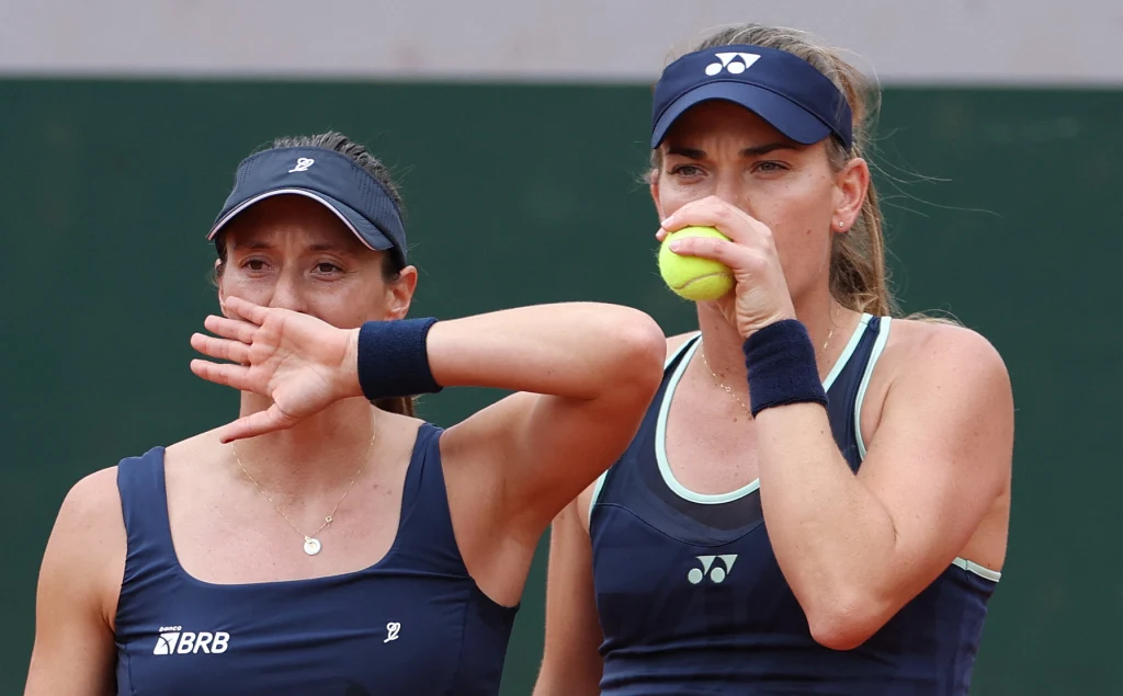 Dwie tenisistki w ciemnych strojach sportowych stoją obok siebie na korcie, jedna trzyma piłkę tenisową przy ustach, druga zasłania usta dłonią, obie koncentrują się przed kolejną akcją.