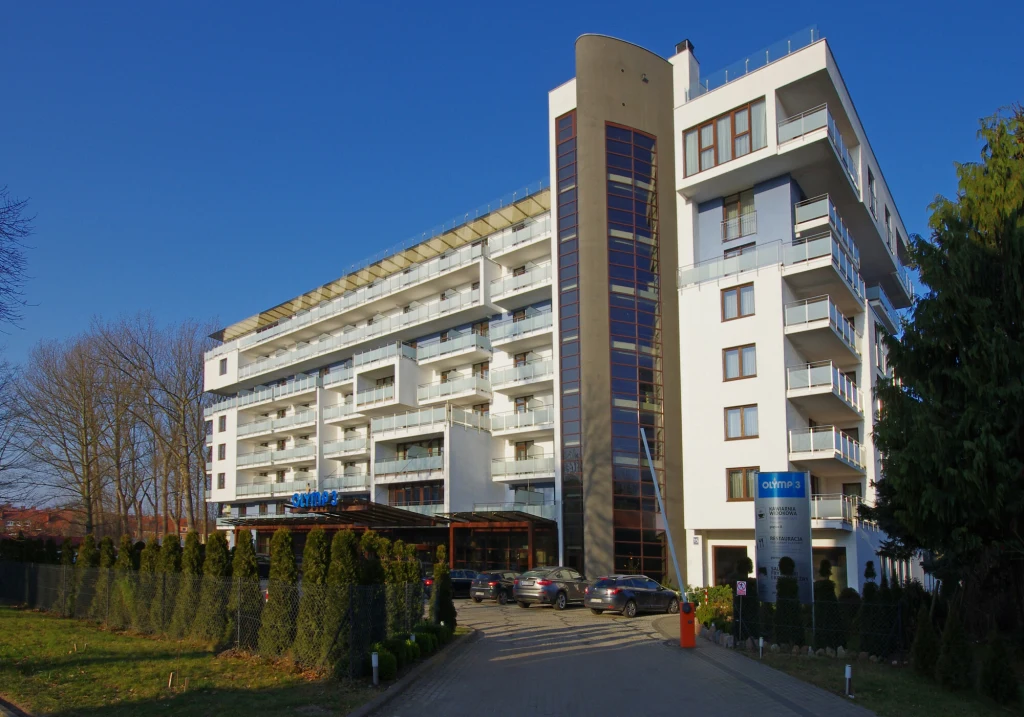 Nowoczesny, wielokondygnacyjny budynek hotelowy z licznymi balkonami i przeszklonymi elewacjami, przed którym zaparkowane są samochody gości, otoczony zadbanym ogrodem i wysokimi drzewami.
