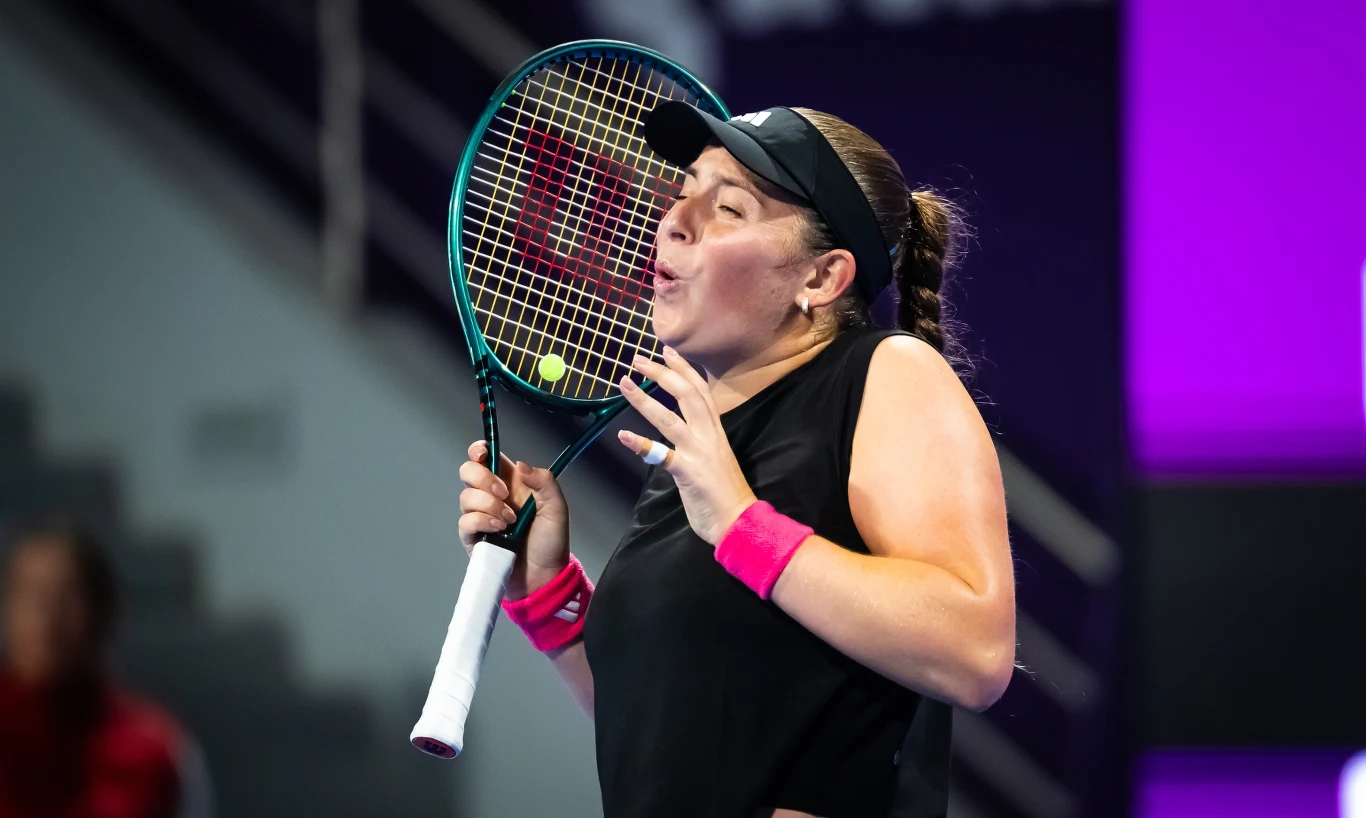 Jelena Ostapenko Tenisistka w czarnym stroju sportowym przygotowuje się do odbicia piłki, trzymając rakietę tuż przy twarzy. Na jej twarzy widoczne skupienie i emocje związane z grą, w tle rozmyte elementy hali sportowej oraz oświetlenie.