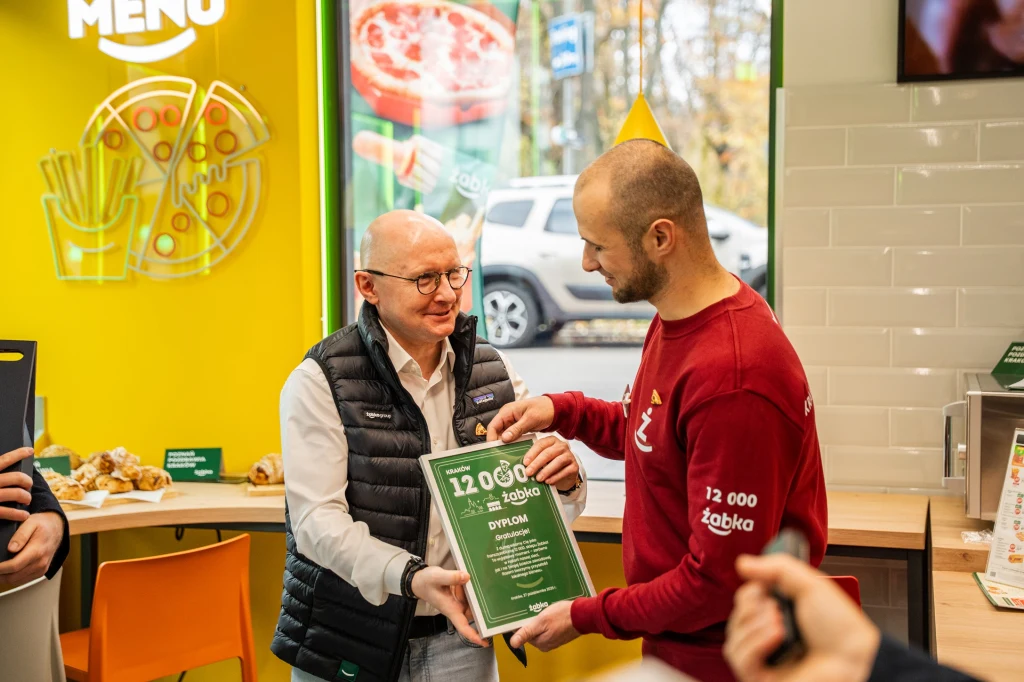 Dwóch mężczyzn wymienia uścisk dłoni i dyplom w nowoczesnym wnętrzu sklepu Żabka, na ścianie widoczne elementy menu i rysunek pizzy, w tle dekoracje oraz fragment ekspozycji produktów spożywczych.