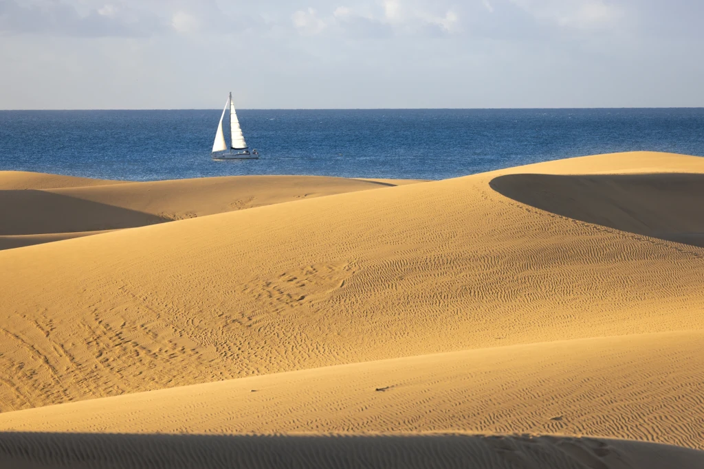 Zachwycający rezerwat ruchomych wydm i kilometry piaszczystych plaż przyciągają na Gran Canarię setki turystów rocznie Jasnożółte wydmy pustynne na pierwszym planie przechodzą w lazurową linię morza, na którym płynie samotny biały żagiel. Lekkie chmury na niebie, kontrast piasku z wodą tworzy spokojny, idylliczny krajobraz.