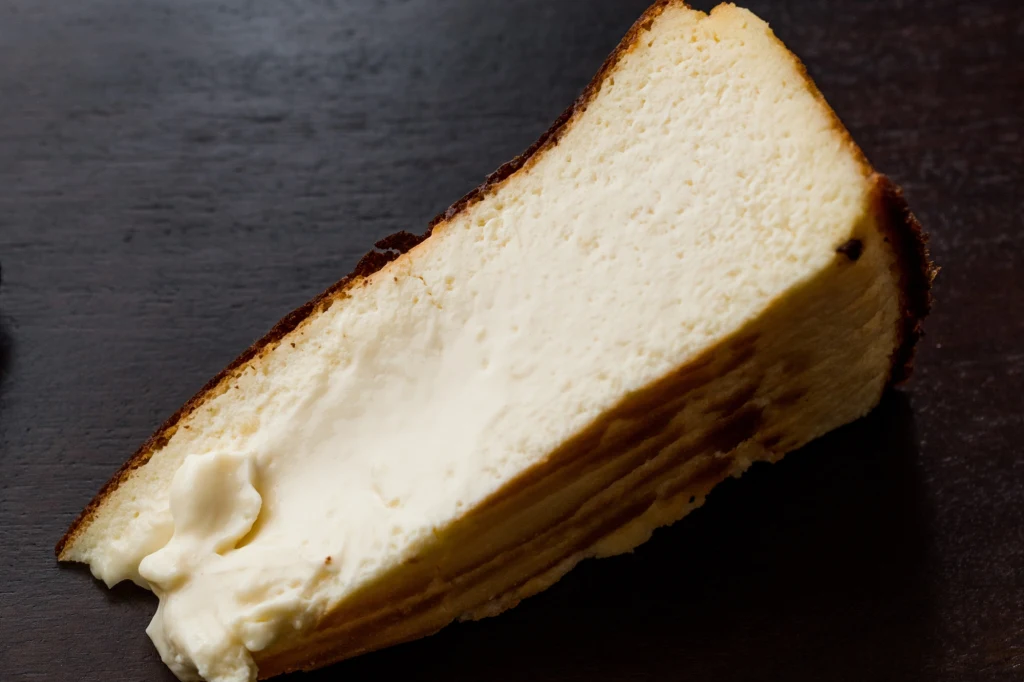 Kawałek jasnego, puszystego sernika z delikatną, przypieczoną skórką na górze, leżący na ciemnym tle.