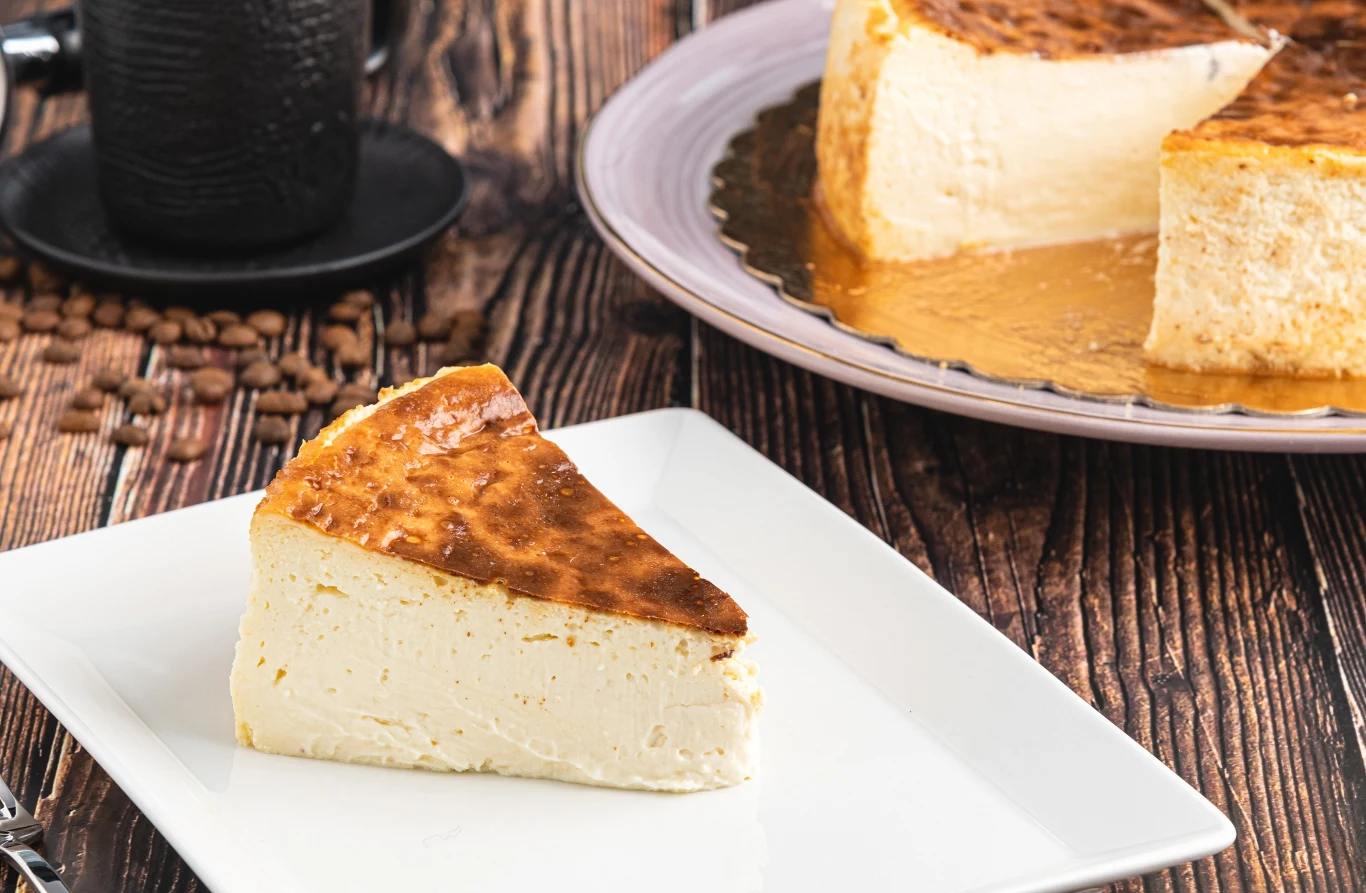 Kawałek sernika o lekko przypieczonej, złocistej powierzchni leży na białym kwadratowym talerzu, obok widoczny jest większy fragment ciasta na okrągłym półmisku ustawionym na drewnianym stole.