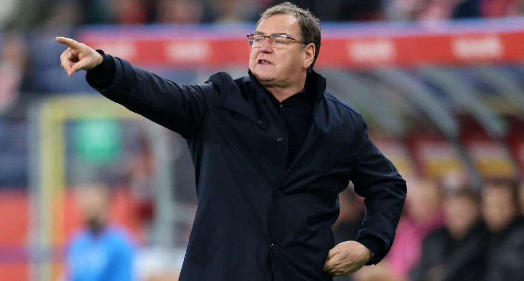 Jan Urban, selekcjoner reprezentacji Polski Mężczyzna w ciemnym płaszczu gestykuluje i wskazuje ręką kierunek, prawdopodobnie znajdując się na stadionie piłkarskim; w tle rozmyte postacie i elementy trybun.