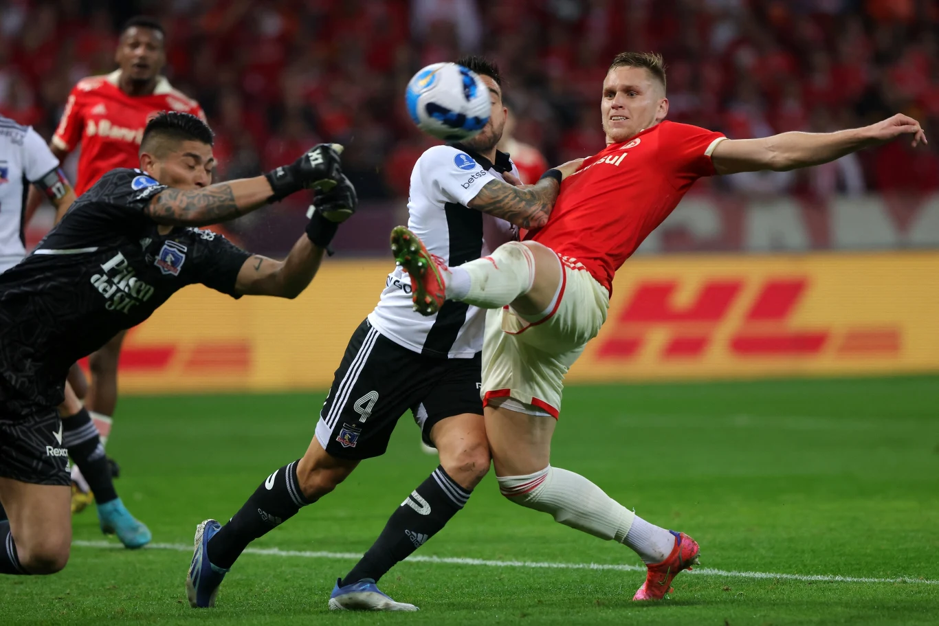 W akcji ofensywnej Alexandre Zurawski (w czerwonej koszulce) Intensywna walka o piłkę podczas meczu piłki nożnej na stadionie, trzech zawodników znajduje się w centrum akcji – bramkarz oraz zawodnik w czarno-białym stroju próbują powstrzymać atakującego w czerwonej koszulce, który wyciąga nogę do piłki.