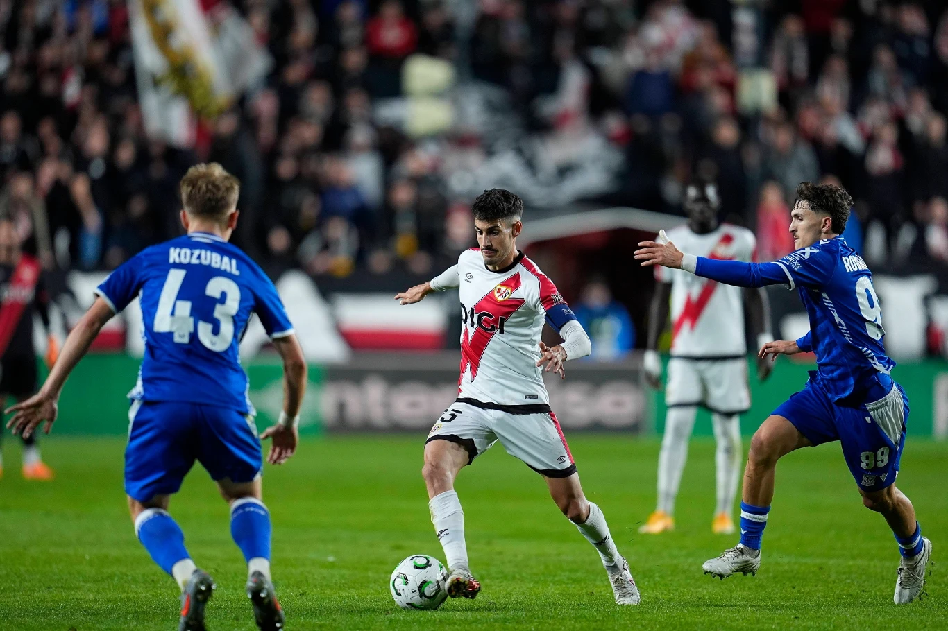 Piłkarz w białym stroju z czerwonym paskiem prowadzi piłkę na murawie podczas meczu, otoczony przez dwóch zawodników drużyny przeciwnej w niebieskich strojach, na boisku widoczna intensywna rywalizacja.