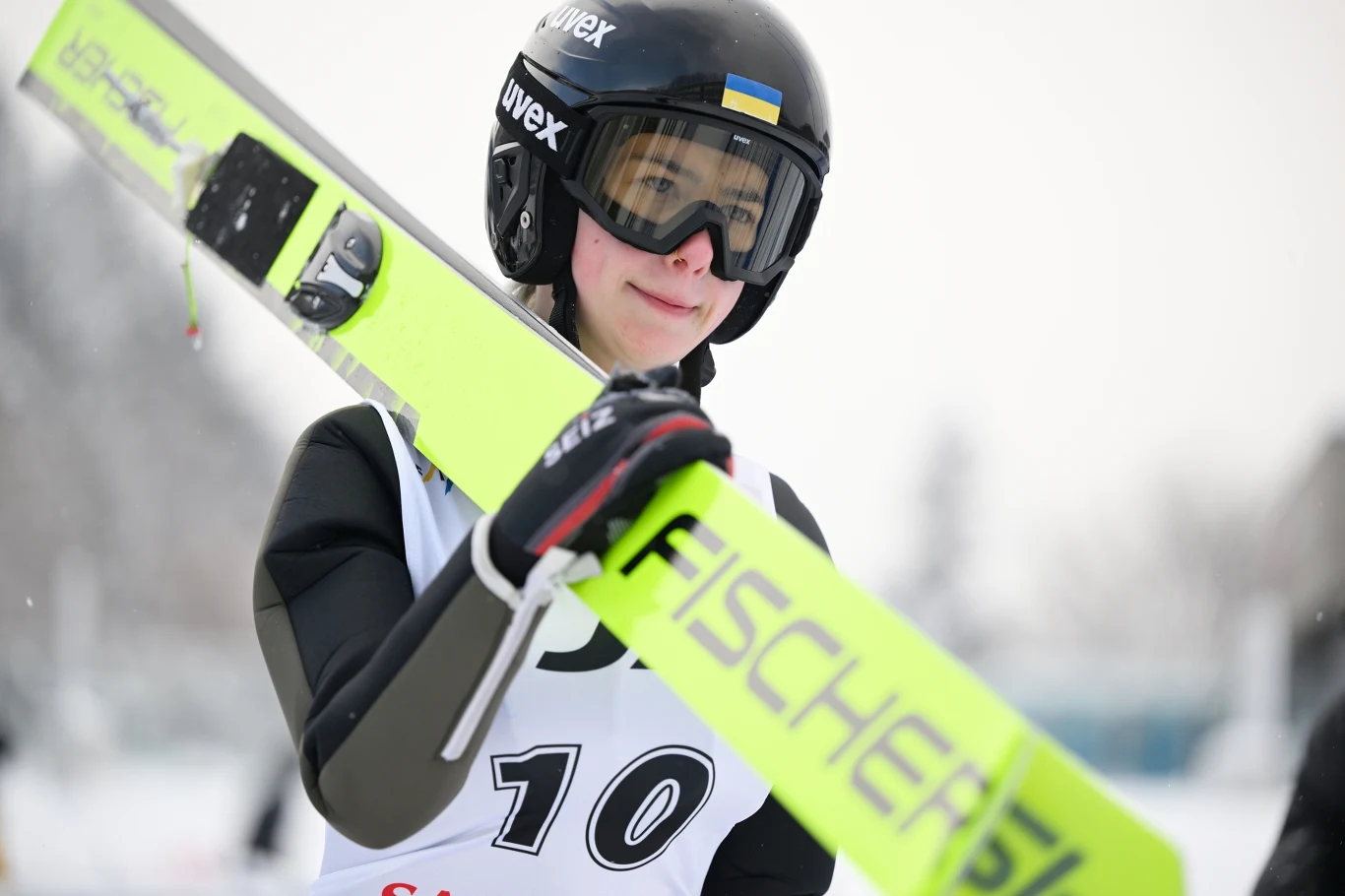 Zawodniczka w stroju narciarskim trzymająca narty skokowe, z numerem startowym i kaskiem, po zakończonej próbie na śnieżnym tle.