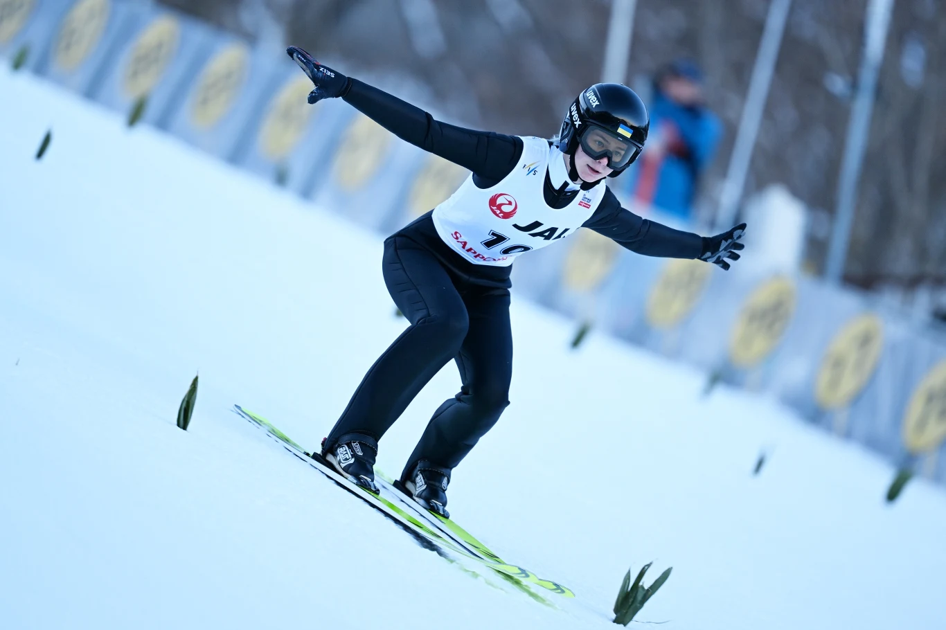 Zawodniczka skacze na nartach, lądując na śniegu podczas zawodów w skokach narciarskich. Ubrana w czarny kombinezon i kask, z rozłożonymi ramionami dla zachowania równowagi. W tle rozmazane bandy oraz ślady po lądowaniach na śniegu.
