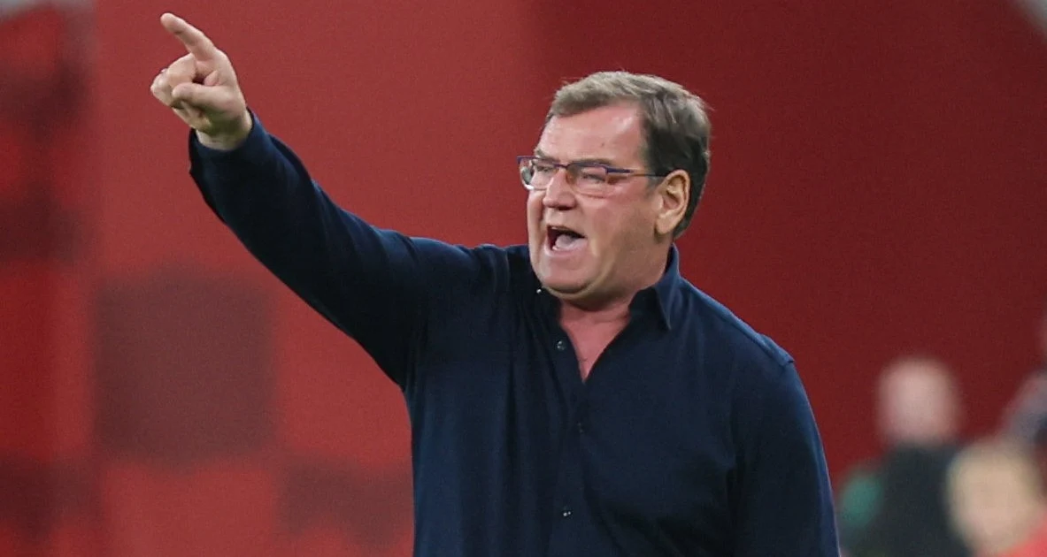 Mężczyzna w okularach, ubrany w ciemną koszulę, energicznie wskazuje palcem w stronę czegoś poza kadrem, gestykulując i wyrażając emocje. Tło jest rozmyte, co skupia uwagę na postaci.