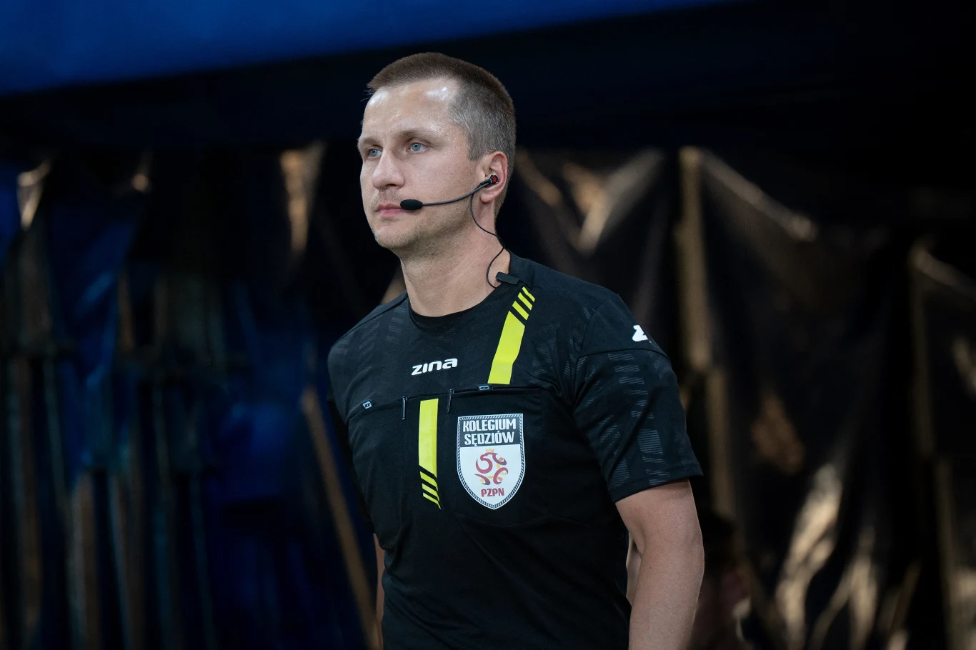 Sędzia piłkarski w czarnej koszulce z herbem Kolegium Sędziów PZPN oraz mikrofonem na głowie stoi na tle ciemnego boiska lub stadionu.