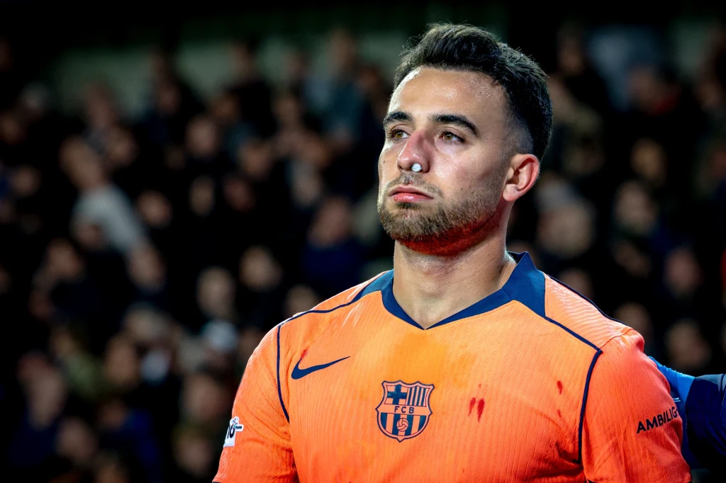Młody piłkarz w pomarańczowej koszulce FC Barcelony z krwią na twarzy i stroju, ze śladami urazu na nosie, stoi na tle nieostrej publiczności, wyraźnie zamyślony lub smutny po meczu.
